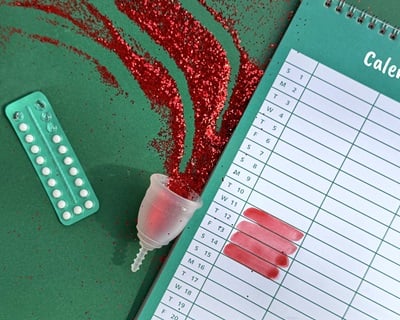 Eine Menstruationstasse mit rotem Glitzer, ein Wecker, Verh&uuml;tungspillen und ein Kalender mit rot markierten Menstruationstagen